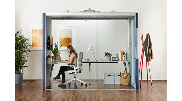 Person working at a desk inside a transparent office pod with plants and decor. rent furniture